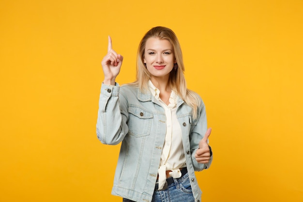 Glimlachende jonge vrouw in denim vrijetijdskleding die wijsvinger omhoog houdt met geweldig nieuw idee, duim opdagen geïsoleerd op geeloranje achtergrond in studio. Mensen levensstijl concept. Bespotten kopie ruimte.