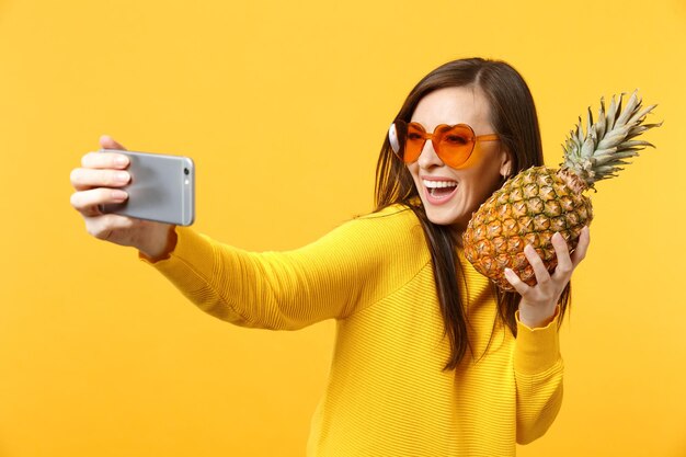 Glimlachende jonge vrouw in hart zonnebril houden ananas fruit, selfie schot op mobiele telefoon geïsoleerd op geeloranje achtergrond doen. Mensen levendige levensstijl ontspannen vakantie concept. Bespotten kopie ruimte