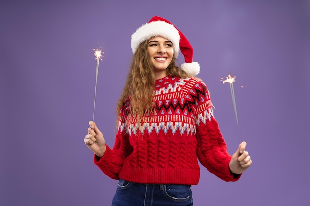 Glimlachende jonge vrouw in kerstmuts danst met brandende sterretjes op paarse studio achtergrond