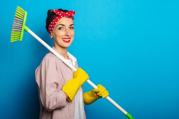 Glimlachende jonge vrouw in schoonmaakhandschoenen die een schoonmaakborstel op blauwe achtergrond houden