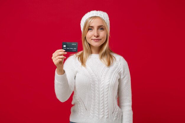 Glimlachende jonge vrouw in witte trui, hoed geïsoleerd op rode muur achtergrond, studio portret. Gezonde mode levensstijl, emoties van mensen, concept van het koude seizoen. Mock up kopie ruimte. Houd een creditcard bij de hand.