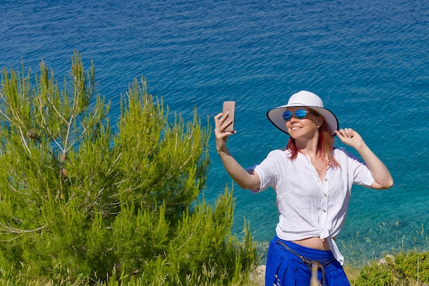 Glimlachende jonge vrouw in zonnebril en strohoed die selfie neemt tegen zee en blauwe lucht