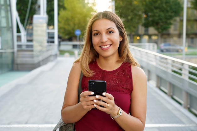 Glimlachende jonge vrouw loopt in de straat met telefoon. Ze houdt gadget in de hand en kijkt naar de camera.