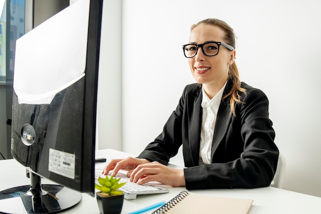 Glimlachende jonge vrouw met een bril werkt zittend op een computer op de werkplek