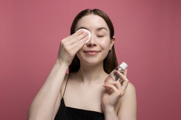 Glimlachende jonge vrouw met gesloten ogen make-up verwijderen met wattenschijfje, fles micellair water op roze achtergrond te houden. Meisje schoonmaak gezicht. Behandeling en cosmetologie. Schoonheidsroutine, huidverzorging