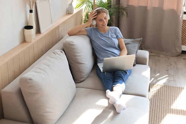 Glimlachende jonge vrouw met koptelefoon en laptop op de bank.