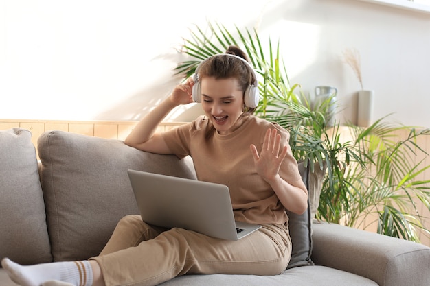 Glimlachende jonge vrouw met koptelefoon en laptop op de bank.