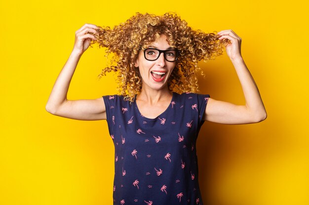 Glimlachende jonge vrouw met krullend haar in een bril met haarlokken in haar handen op gele achtergrond