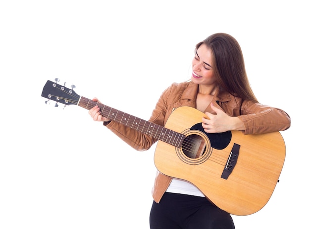 Glimlachende jonge vrouw met lang haar in bruine jas en zwarte broek die een gitaar vasthoudt en speelt