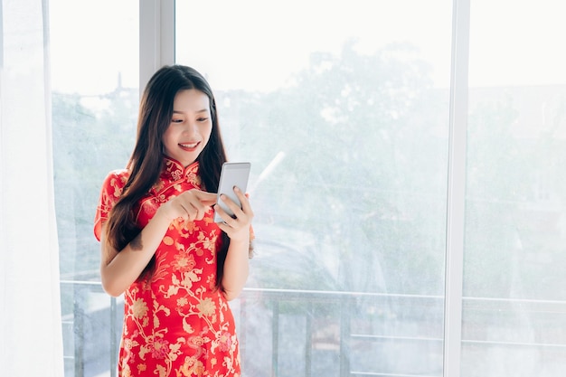 Glimlachende jonge vrouw met mobiele telefoon terwijl ze bij het raam thuis staat