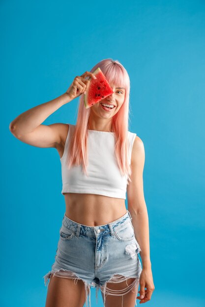 Glimlachende jonge vrouw met roze haar die een plakje watermeloen boven haar oog houdt terwijl ze geïsoleerd staat