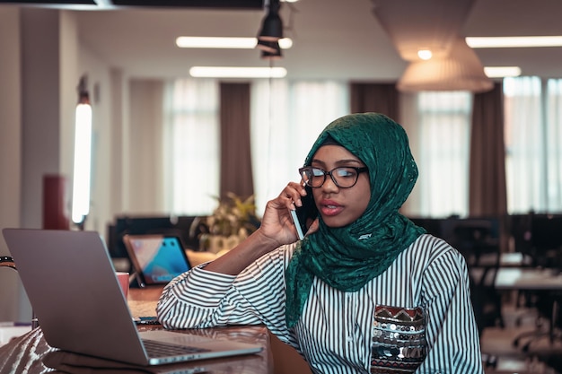 Glimlachende jonge vrouw praten over smartphone werk op laptop kijken in afstand denken. Gelukkig Afro-Amerikaanse klant klant met behulp van moderne gadgets mobiele telefoon en computer op het kantoor aan huis. Foto's van hoge kwaliteit