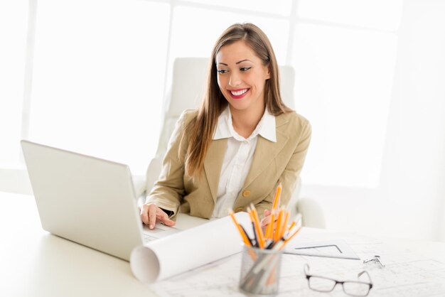 Glimlachende jonge vrouwelijke ingenieur die bij laptop in haar bureau werkt.