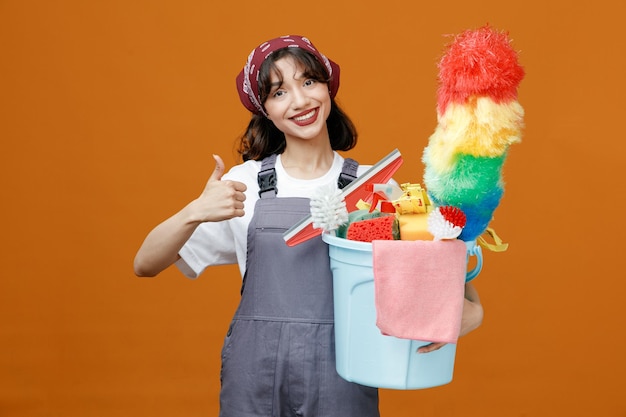 Glimlachende jonge vrouwelijke schoonmaker die uniform en bandana draagt die een emmer schoonmaakgereedschap vasthoudt en naar een camera kijkt die duim omhoog laat zien geïsoleerd op een oranje achtergrond