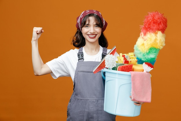 Glimlachende jonge vrouwelijke schoonmaker die uniform en bandana draagt met emmer schoonmaakhulpmiddelen die naar de camera kijken en een sterk gebaar maken dat op een oranje achtergrond wordt geïsoleerd