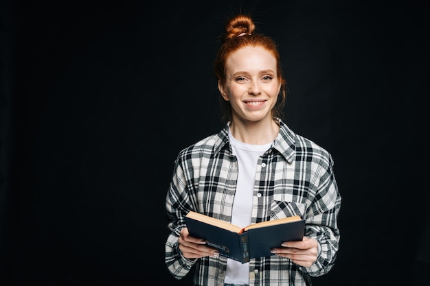 Glimlachende jonge vrouwelijke student die een geopend boek vasthoudt en naar de camera kijkt op zwarte achtergrond