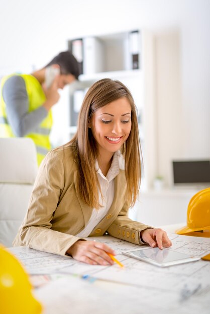 Foto glimlachende jonge vrouwenconstructeur die blauwdruk op tablet analyseert bij bureau in bureau.