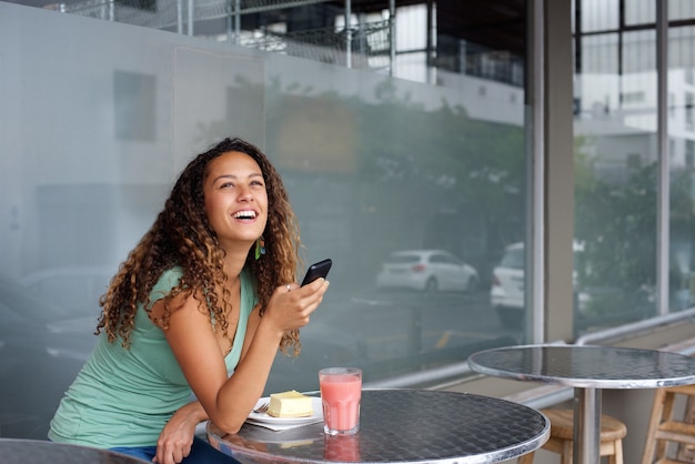 Glimlachende jonge vrouwenzitting bij een koffie die mobiele telefoon met behulp van