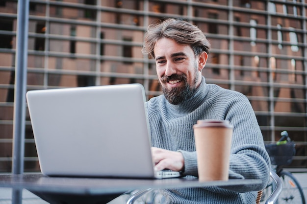 Glimlachende jonge zakenman die laptop gebruikt om op afstand te werken vanaf het koffieterras Portret van een succesvolle freelancer of student die aan een nieuw project werkt Bedrijfsconcept