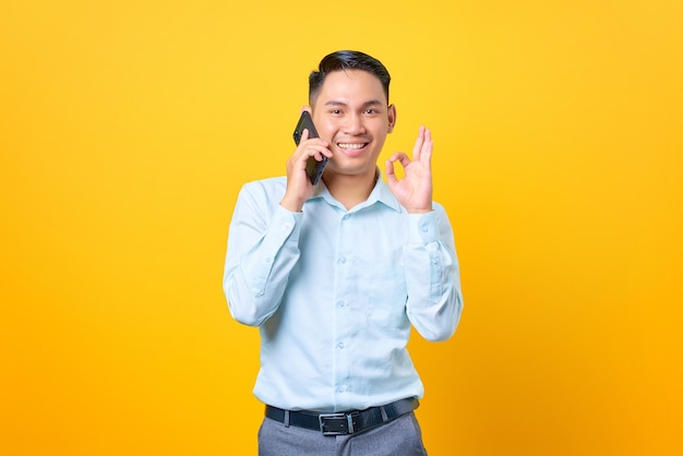Glimlachende jonge zakenman die op een smartphone spreekt en goed gebaar doet op gele achtergrond