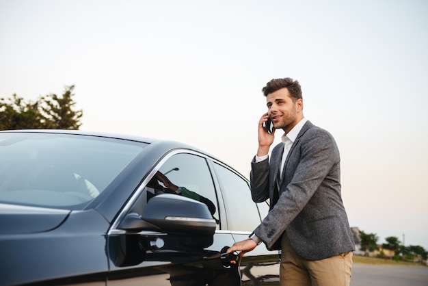 Glimlachende jonge zakenman in kostuum dat zich bij zijn auto bevindt