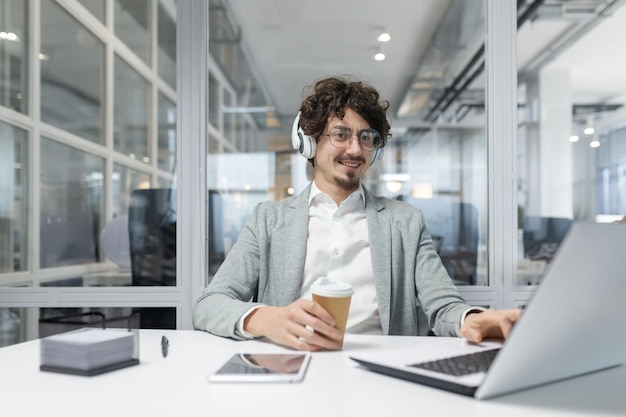 Glimlachende jonge zakenman met krullend haar en koptelefoon die aan een laptop werkt in een goed verlichte kantoor