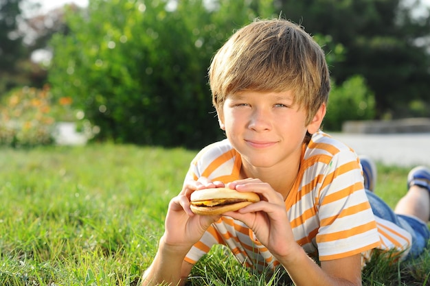 Glimlachende jongen die op groen gras in park ligt dat hamburger in openlucht eet. Ongezond fastfood