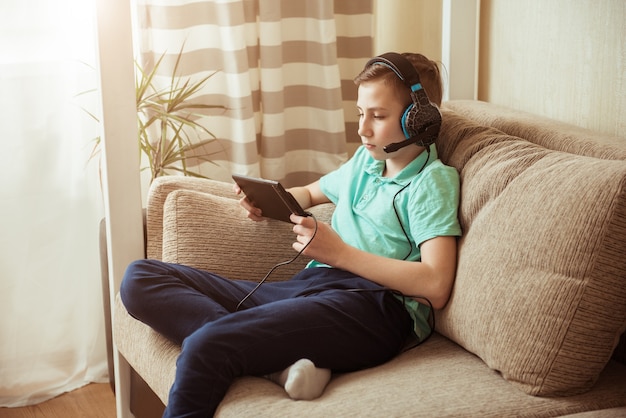 Glimlachende jongen doet zijn huiswerk in hoofdtelefoons en met een tablet. Afstandsonderwijs in quarantaine