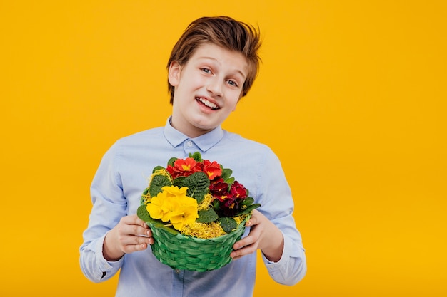 Glimlachende jongen met mand met bloemen in zijn hand, in het blauwe overhemd, dat op gele muur wordt geïsoleerd