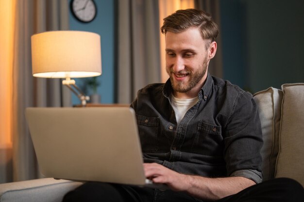 Glimlachende jongen werkt op afstand in zijn eigen woonkamer op zijn bank houdt zijn laptop op schoot