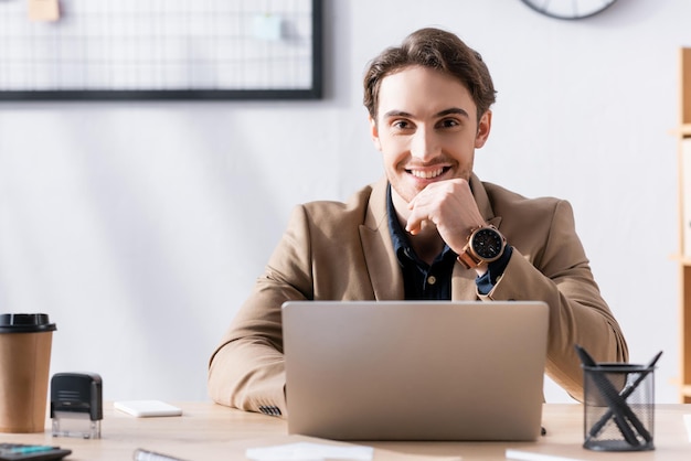 Glimlachende kantoormedewerker die naar de camera kijkt terwijl hij in de buurt van een laptop aan een bureau in kantoor zit op wazig