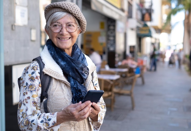 Glimlachende kaukasische senior vrouw die in een stadsstraat loopt met een mobiele telefoon die positiviteit uitdrukt, een goed humeur met een rugzak