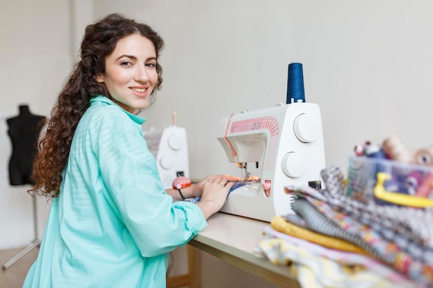 Glimlachende kleermaker met donker krullend haar in een kleurrijk shirt dat vrolijk in de camera kijkt terwijl hij met de naaimachine werkt in een moderne naaiatelier