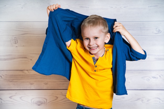 Glimlachende kleine jongen in gele t-shirt die zijn blauw overhemd opneemt. portret van een vrolijk kind heeft plezier