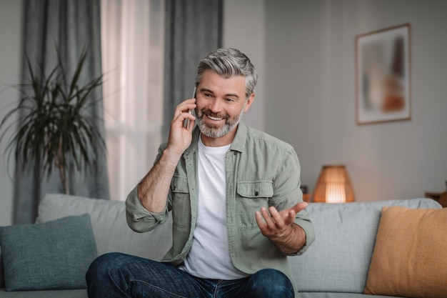 Glimlachende knappe gepensioneerde europese man met baard belt door smartphone praten op afstand in woonkamer interieur