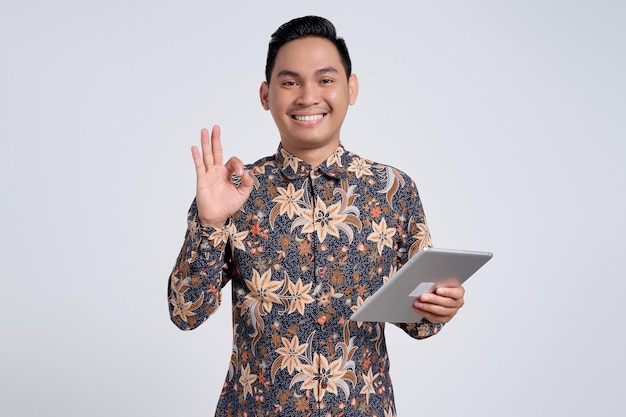 Foto glimlachende knappe jonge aziatische man in een batik shirt met een digitale tablet en een ok-teken geïsoleerd op een witte achtergrond mensen levensstijlconcept