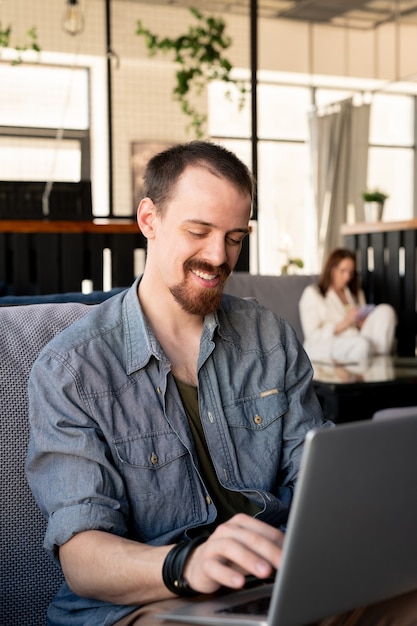 Glimlachende knappe jonge bebaarde man in denim overhemd typen op moderne laptop tijdens het werken in café