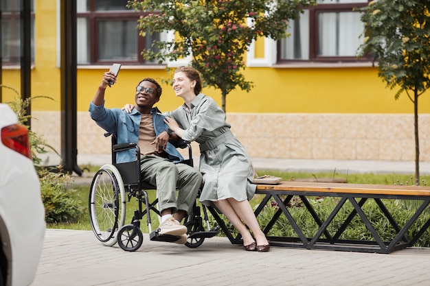 Glimlachende knappe jonge gehandicapte Afro-Amerikaanse man met een bril die in een rolstoel zit en een selfie neemt met zijn vriendin buitenshuis