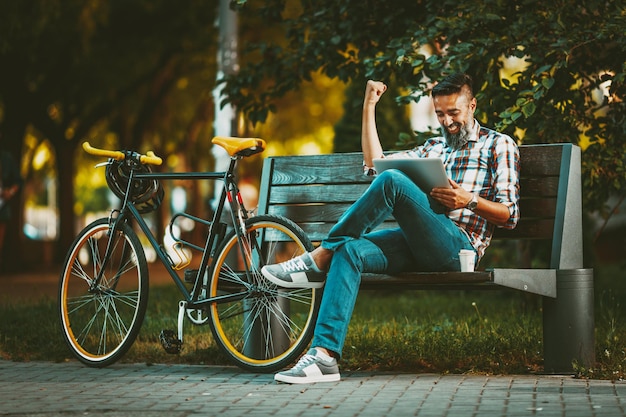 Glimlachende knappe jonge man is op een pauze. Hij zit op een bank en werkt aan laptop, succes vierend, naast de bank rust een fiets.