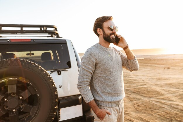 Glimlachende knappe jongeman die mobiele telefoon gebruikt terwijl hij bij de auto op het zonnige strand staat en belt