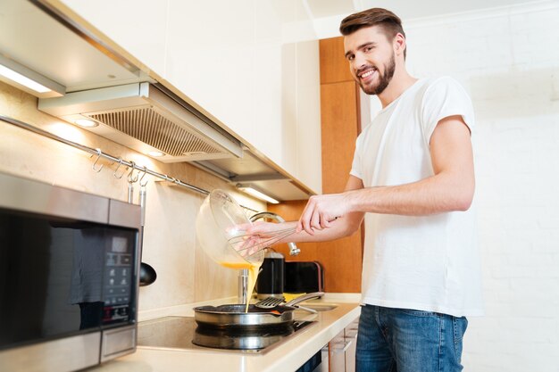 Glimlachende knappe jongeman die staat en omelet kookt voor het ontbijt