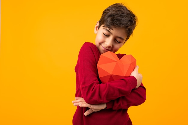 Glimlachende knappe jongen met papieren hart voor valentijnsdag op oranje achtergrond.