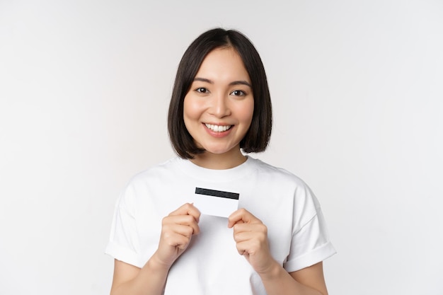 Glimlachende Koreaanse vrouw die creditcard met gelukkig gezicht toont die zich in t-shirt over witte achtergrond bevinden