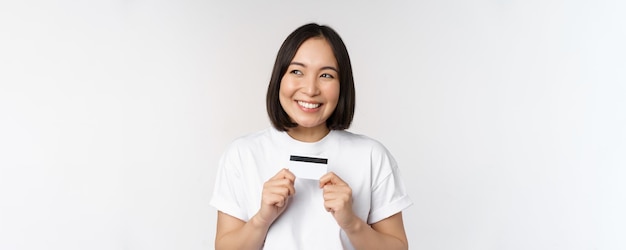 Glimlachende Koreaanse vrouw die creditcard met gelukkig gezicht toont die zich in t-shirt over witte achtergrond bevinden