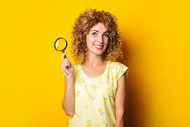 Glimlachende krullende jonge vrouw met een vergrootglas op een geel oppervlak