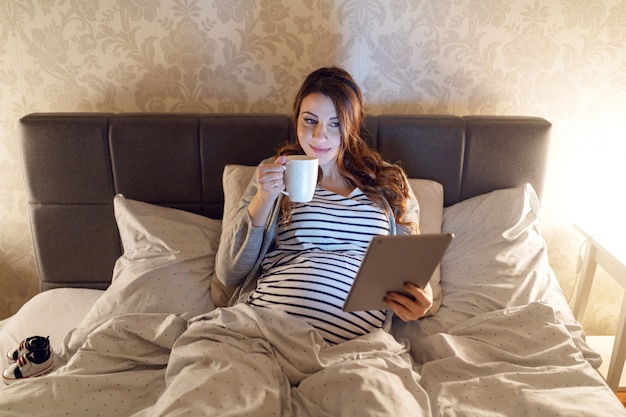 Glimlachende leuke zwangere blanke vrouw met lang bruin haar liggend in het bed, thee drinken en lezen over baby's op tablet. Avond tijd.