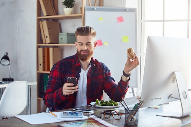 Glimlachende man die hamburger eet op het werk tijdens het gebruik van de telefoon