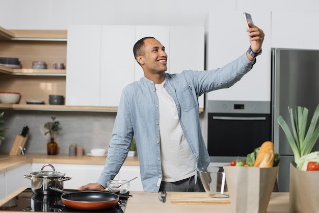 Glimlachende man die selfie doet of een videogesprek voert terwijl hij gezond veganistisch eten kookt in het keukeninterieur