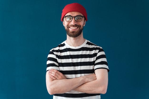 Glimlachende man in glazen en rode hoed op een blauwe achtergrond. Brunette man geïsoleerd op blauwe achtergrond gekruiste armen