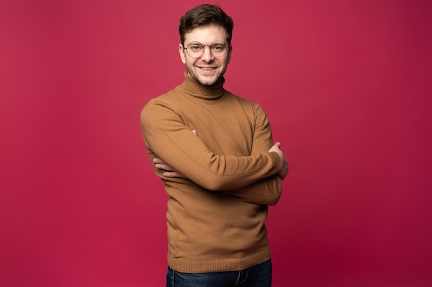 Glimlachende Man in glazen en trui staan met gekruiste armen en wegkijken. Geïsoleerde roze achtergrond.